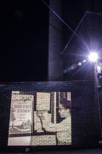 Baden Street Settlement at night with projected image of woman and poster that reads: "Come to the Baden Street Settlement: clubs, teams, dances, good times for everybody."