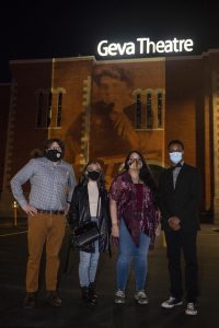 Professor Joseph Scanlon and three students standing in front of the Geva Theatre with a projected image of Emma Goldman.