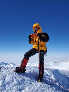 Ilina Arsova standing in winter mountaineering gear atop a snowy mountain holding a small flag of North Macedonia