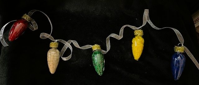 Decorative garland featuring five large, bulb-shaped ornaments arranged horizontally against a black background. The ornaments are attached to a sheer, white ribbon that loops and curls between each bulb, creating a festive and elegant look. The bulbs resemble oversized holiday light bulbs each filled with a different colored yarn.