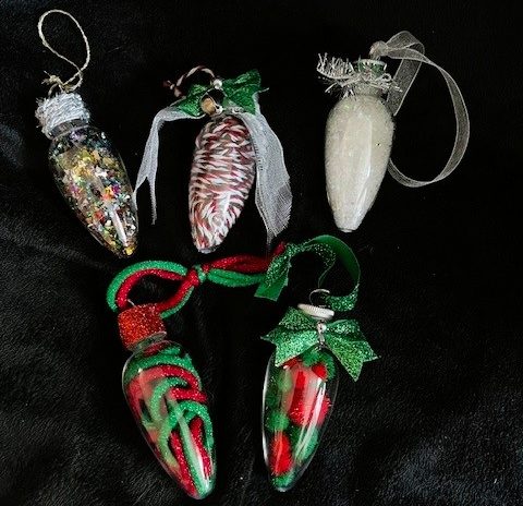 The image shows five decorative ornaments shaped like oversized holiday light bulbs, arranged on a black background. The ornaments are positioned in two rows: three at the top and two at the bottom. Each ornament is unique, filled with different festive materials and adorned with decorative caps and ribbons.
