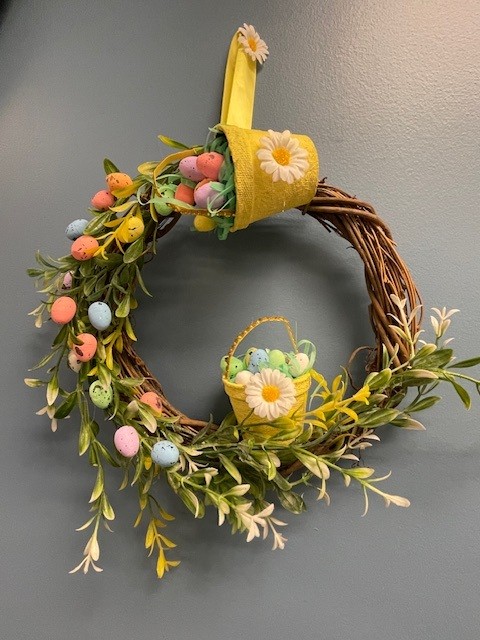 An Easter‑themed wreath made from a natural, vine‑style base. It is embellished with soft green foliage that circles around the bottom half. Among the greenery are small, pastel-colored decorative eggs in shades of pink, blue, yellow, green, and orange, each with tiny speckles for added detail.