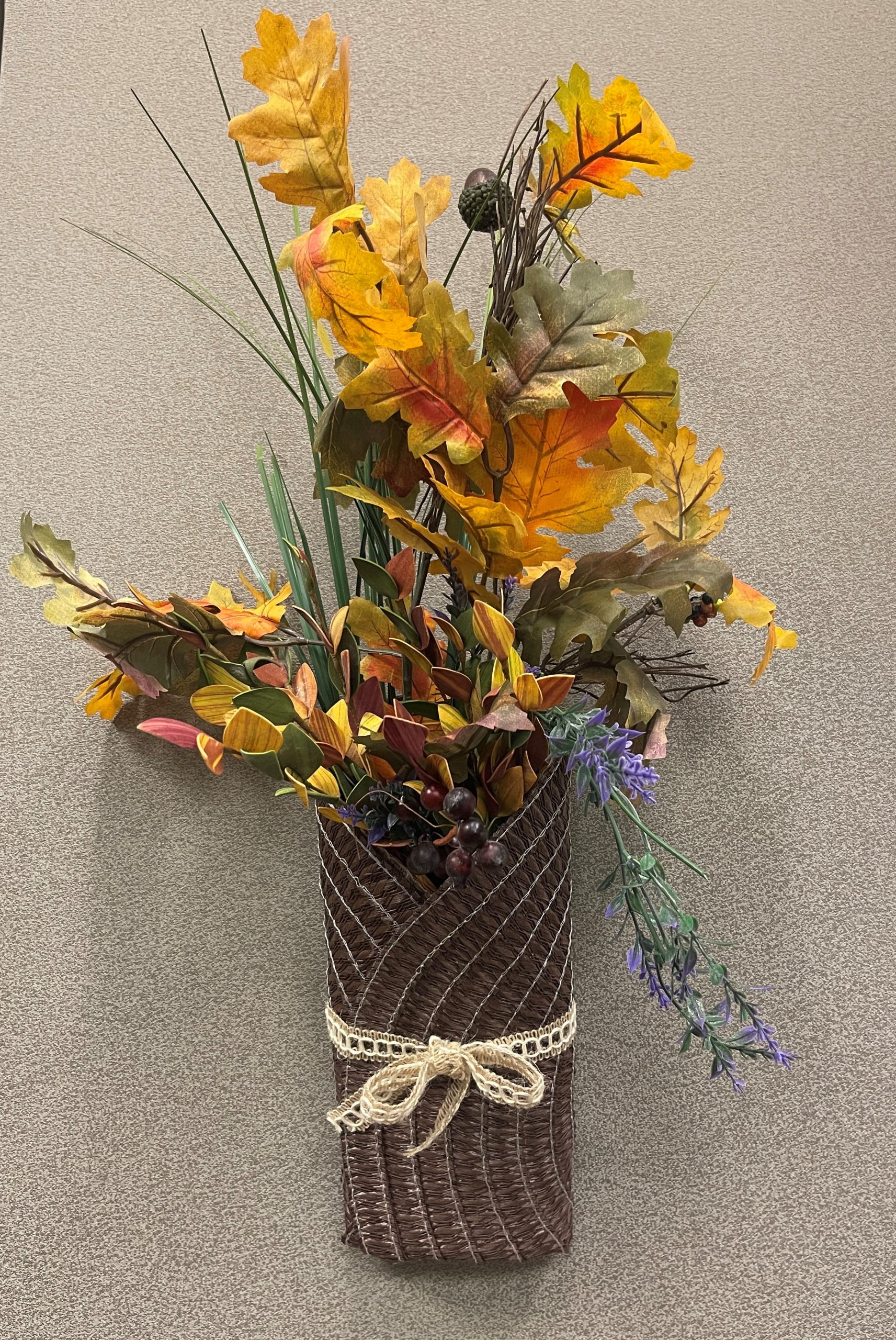 The wall decor is composed of artificial fall foliage in shades of gold, orange, rust, burgundy, green, and brown. Several large maple style leaves extend upward, creating height and a cascading, natural appearance. Tall, narrow green grass stems rise vertically through the center of the arrangement. Mixed throughout the design are small berry clusters in dark red and burgundy tones, pinecone accents, variegated leaves in autumn colors, sprigs of purple lavender-like flowers, greenery and trailing stems that extend outward from the container. The arrangement is housed in a tall rectangular dark brown vase made from a folded round placemat. Decorative white lace trim is wrapped around the center and tied into a small bow on the front.