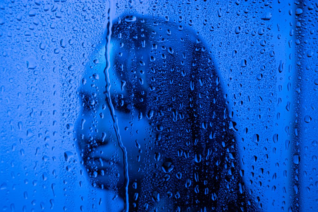 Photograph of a side view of a woman's face behind a glass pane with water droplets and the entire photograph has a blue hue.
