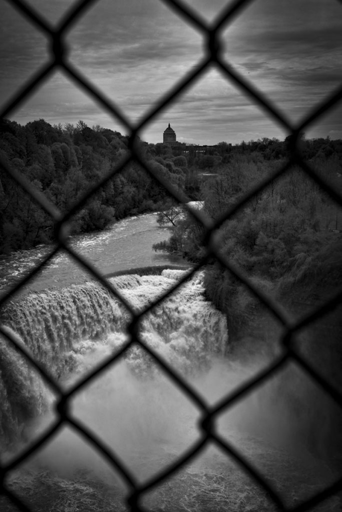 A black and white photograph taken through a chain link fence, in the foreground in a waterfall, and in the background is the Rochester skyline and trees.