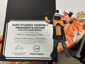 Tiberius with Student Assembly President’s Award: Legal-sized paper cutout of Tiberius, the MCC mascot, stands next to the paper certificate of the SUNY Student Assembly President’s Award.