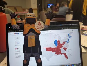 A legal-size paper cutout of Tiberius, the MCC mascot, is partially in front of a laptop computer screen showing the electoral map with results. 