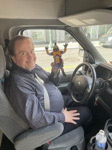 Tiberius and Bus Driver SGA Trip – Joseph Scanlon, Ph.D., an associate professor of political science, is shown seated in the driver's seat inside of the MCC bus, smiling at the camera and holding up the paper cutout Tiberius.