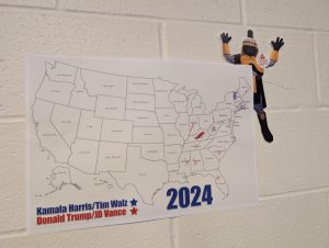 A legal-size paper cutout of Tiberius, the MCC mascot, is half hidden behind a large United States map on the wall, with the outline of states showing which candidates are winning which states. 