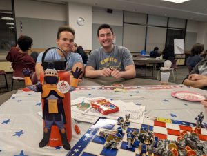 A legal-size paper cutout of Tiberius, the MCC mascot, is leaning against a large water bottle on a table with a chess-like game board and an Uno box. Two students are in the background, smiling at the table. 