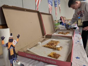 A legal-size paper cutout of Tiberius, the MCC mascot, is to the left of the photo against a roll of paper towels with large boxes of pizza next to him on the table. 