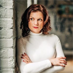 Professional headshot of Danielle Froment wearing a white shirt and leaning against a wall with her arms crossed.