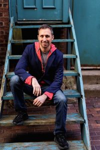 Tony Leuzzi sitting outside on older, light blue wooden stairs smiling directly at the camera and wearing casual clothing.