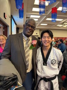 Dr. Calvin Gantt standing on the left with a brown suit jacket and light button down shirt with his arm around Matthew Ra, dressed in a traditional Taekwondo uniform called a dobok.