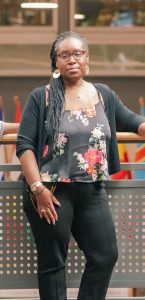 Yolanda Johnson is standing in front of a metal fence with her arm on a railing, wearing black pants, a black tank top with red and white flowers, and a black cardigan. 