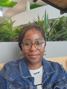 Headshot of Kim DeLardge smiling at the camera wearing a denim jacket and white shirt. Plants are visible in the background.
