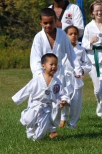 Matthew Ra, as a toddler, running in the grass dressed in a traditional Taekwondo uniform called a dobok. There are other children in doboks running behind him. 