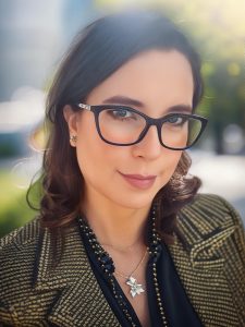 Headshot of Antonia Custodio outside, smiling at the camera. She is wearing glasses, a dark shirt, and a blazer.