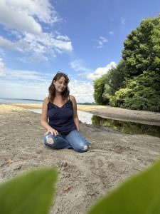 Maria Brandt sitting on the sand in casual attire with water and trees in the background.