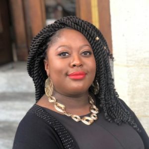 A professional headshot of Afiya Farrell wearing a black top and gold jewelry as she smiles at the camera.