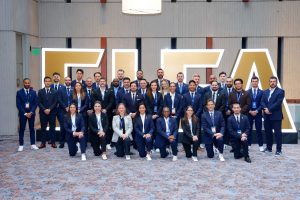Large group of team liaisons wearing matching dark suits in front of FIFA sign.
