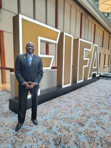Kevin Messam wearing a dark suit is standing next to a large FIFA sign.