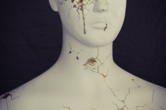A ceramic sculpture of a bust and upper body of a male figure , th efigure is all smooth white, but cracks in the face have been repaired with a gold substance. The chest area has a large hole in it, and gold liquid is on the edge of the hole, and is starting to close the hole.