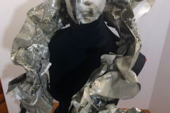 A sculpture of an Elizabethan collar and a face mask both supported by a black sewing manikin. Both the collar and mask are the color of melting snow on the roadside (marbleized grays, browns, and white). The collar is ruffled in the style of queen Elizabeth. The entire outfit is made of paint that after being allowed to dry in flat pools, was peeled up and then sewn into these forms. (Left View)