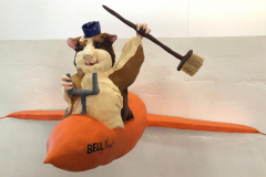 A sculpture of a guinea pig flying a bright orange plane, it wears a small hat and holds a broom above his head. (Straight on view)