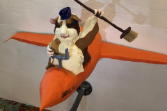 A sculpture of a guinea pig flying a bright orange plane, it wears a small hat and holds a broom above his head. (Slightly elevated view)
