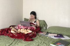 A photograph of a young woman sitting up in a bed, a computer is on her lap and there are books spread out on her left. She looks intently at her computer.