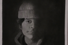 Charcoal drawing of a young man wearing a knit cap. The drawing has extremely high contrast with harsh light on his right cheek, leaving his left side in deep shadow.