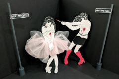 A dimensional illustration. Two paper-doll figures of the same eyepatch-clad, curly-haired young woman are set against a three-dimensional black backdrop. Two street signs create an intersection that frames the figures in the composition. The signs themselves frame the scene with a caption that reads “Then I am only…what I made myself.” The rightmost figure, dressed in a black vinyl skirt and tall pink boots, appears to be stabbing her counterpart on the left. The left figure stares on, seemingly unaware of the wound seeping through her pale pink and white dress. The same wound is present on the stabbing figure, denoting that the two women are the same person. (Full view)