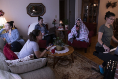 A photograph showing a living room occupied by five different versions of the same woman, each doing a different task. One sits with a cup of tea looking out the window, one sits on the couch with a tv remote in her hand, one sits in a chair working on a computer, one sits in a different chair with a blanket over her head, and the final one sits at a piano playing it.