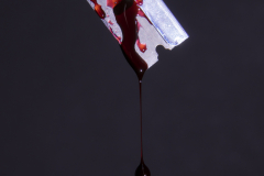 A photograph of a razor blade hovering in air against a dark background, and it has some blood on it and it is slowly dripping off the lowest point of the rzor blade.