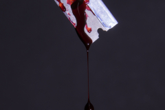 A photograph of a razor blade hovering in air against a dark background, and it has some blood on it and it is slowly dripping off the lowest point of the rzor blade.