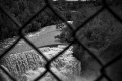 A black and white photograph taken through a chain link fence, in the foreground in a waterfall, and in the background is the Rochester skyline and trees.