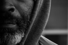 A black and white photograph showing half of a mans face, wearing a hoodie. He has some grey in his beard, and he looks intently at the camera.
