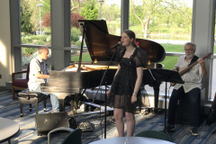 This is a close up photograph of the three musicians playing music at the opening reception. On the left is a piano player, in the center a woman sings, and on the right a man plays the bass guitar.