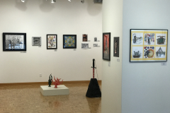 This is a photograph of the 44th annual student art exhibition installation. In this image we are seeing the rear wall of the gallery with 13 pieces of art installed in the space.