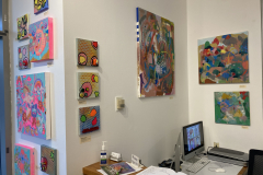 Exhibition installation of desk area containing many small colorful paintings.