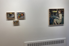 Installation shot of the exhibition, with three small paintings of various fruit and seeds are hanging on the left, and a painting of a woman sitting at a bar s hanging on the right.