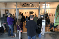 An image taken at the opening while the two artists are giving a gallery lecture. The artists are in the back of the gallery and many people are inside the gallery, standing and on chairs, listening to the artists.