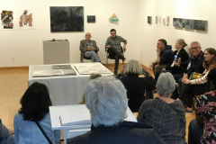 An image taken at the opening while the two artists are giving a gallery lecture. The artists are in the back of the gallery and many people are inside the gallery, standing and on chairs, listening to the artists.