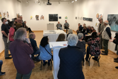 An image taken at the opening while the two artists are giving a gallery lecture. The artists are in the back of the gallery and many people are inside the gallery, standing and on chairs, listening to the artists.