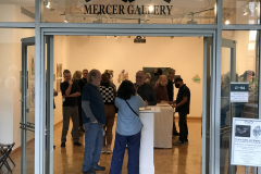 An image from the opening reception looking into the gallery from outside, many people are standing around looking at the artwork inside the gallery.