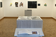 This is a photograph showing the inside of the Mercer Gallery. We see a variety of artwork hanging on the walls and in the center of the image we see pedestals containing various artist books.