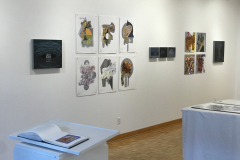 This is a photograph showing the inside of the Mercer Gallery. On the wall hangs various artwork including paintings and drawings, and in the foreground there are two pedestals containing artist books with more artwork in them.
