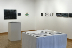 This is a photograph showing the inside of the Mercer Gallery. On the wall hangs various artwork including paintings and drawings, and in the foreground there are two pedestals containing artist books with more artwork in them.