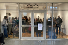 An image from outside the gallery, and inside are a group of people listening to the artist speak.